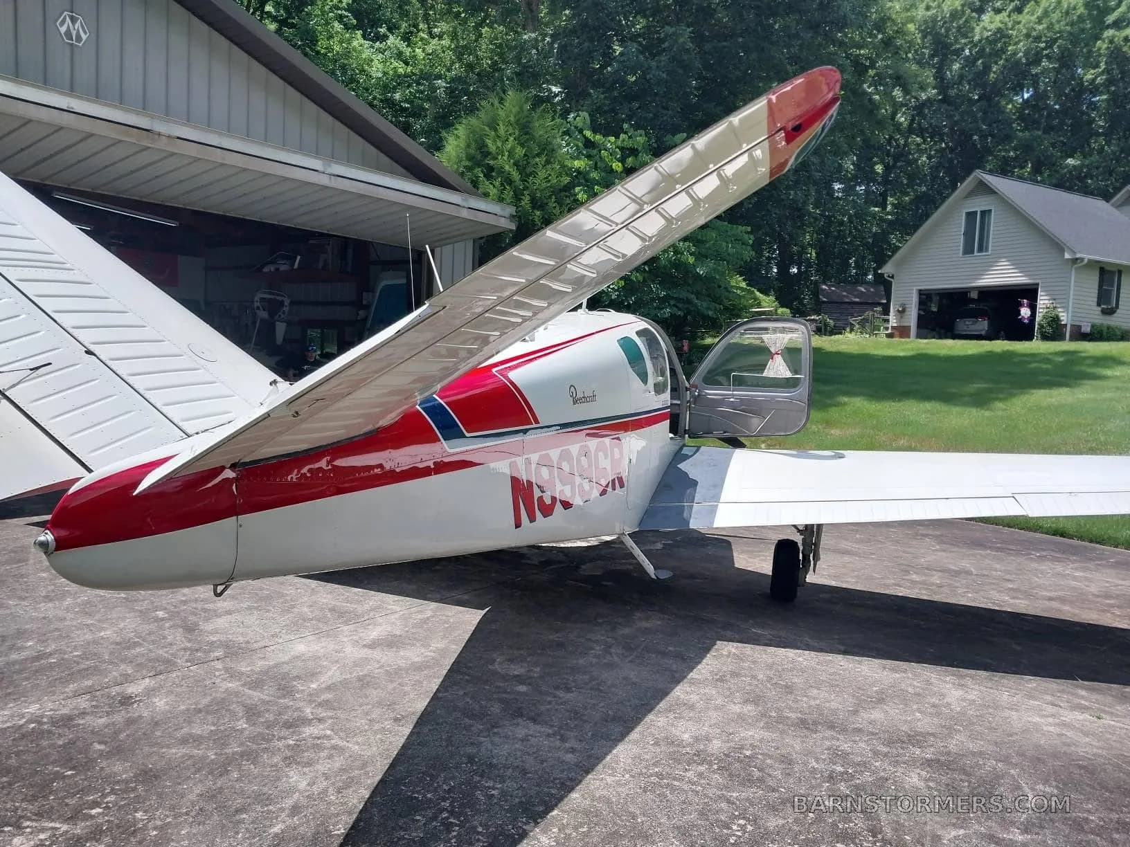 A white Beechcraft Bonanza with red and blue stripes, registration N999CR, viewed from the rear, parked next to a hangar.