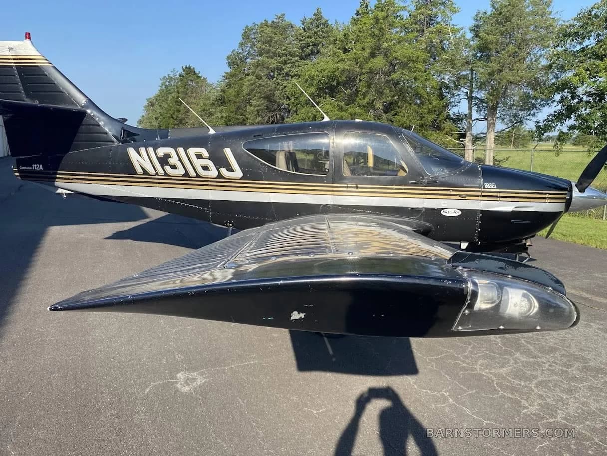 A black Cessna 112A aircraft with gold and silver stripes, registration N316J, parked on asphalt with trees in the background.
