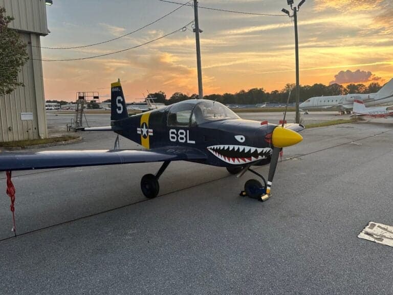 A blue and yellow aircraft with shark teeth painted on the nose is parked on the tarmac at sunset.