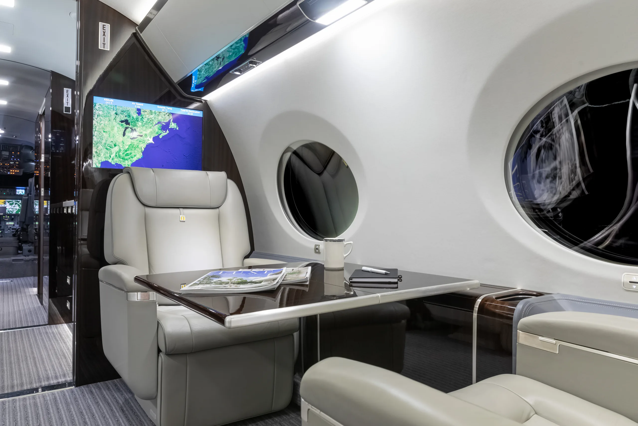 A close-up of a light grey leather seat and a dark wood table in the private jet cabin, with a map displayed on a wall-mounted screen.