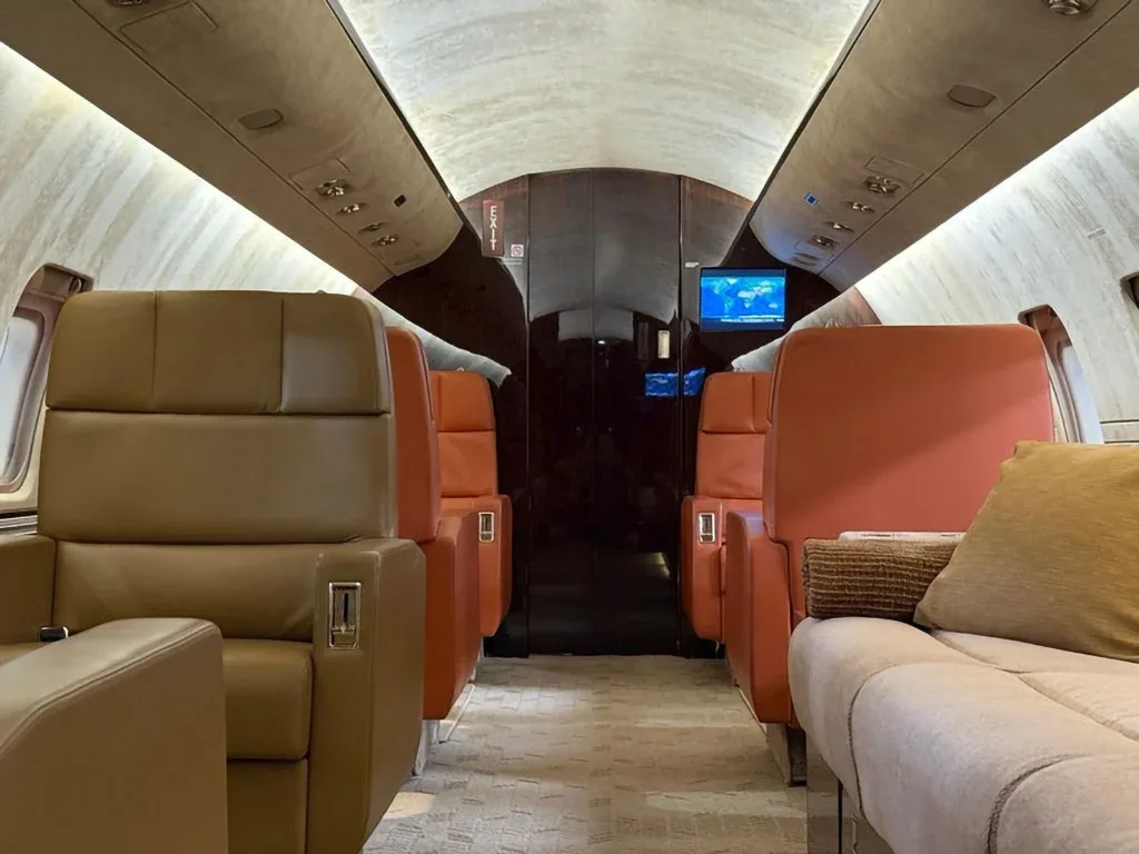 The private jet cabin interior shows a mix of beige and orange leather seats, with a screen displaying a map at the rear.