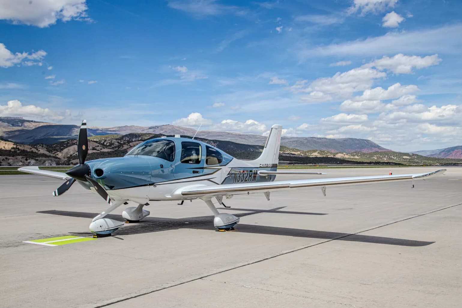 A blue and white Cirrus SR22T GTS aircraft, registration N332RM, parked on an airport tarmac with mountains in the background.