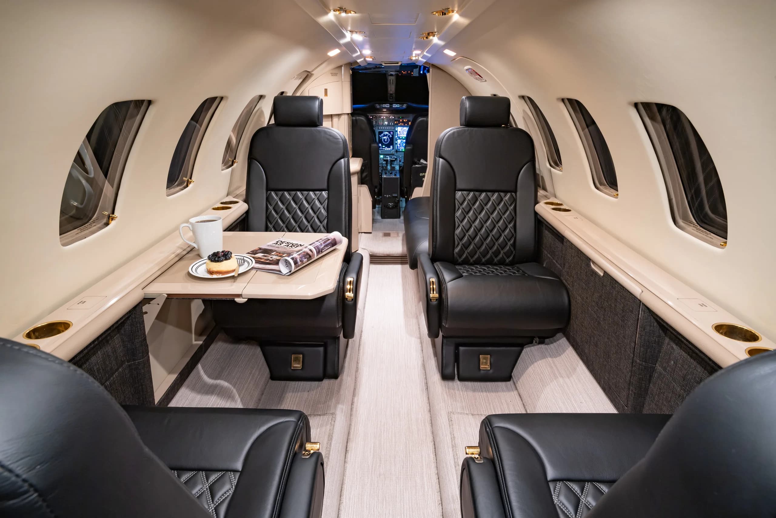 View from the rear of the aircraft cabin looking forward, showing black quilted leather seats, a table with refreshments, and a partial view of the cockpit.