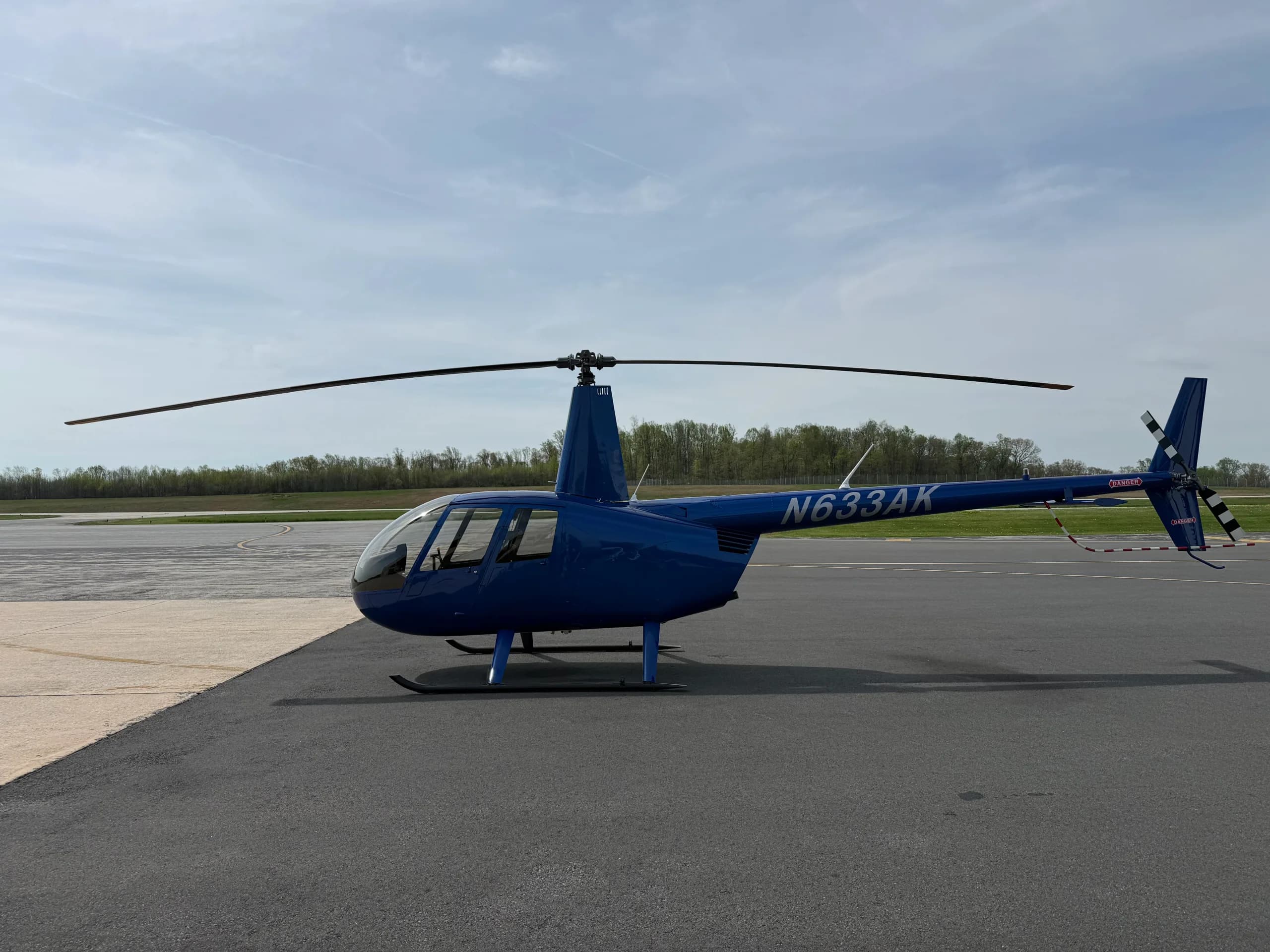 A blue Robinson R44 helicopter, N633AK, parked on an asphalt tarmac with a distant tree line.