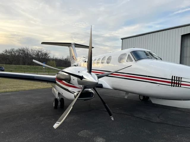A close-up front-right view of the propeller and nose of a white Beechcraft King Air with red and black stripes.