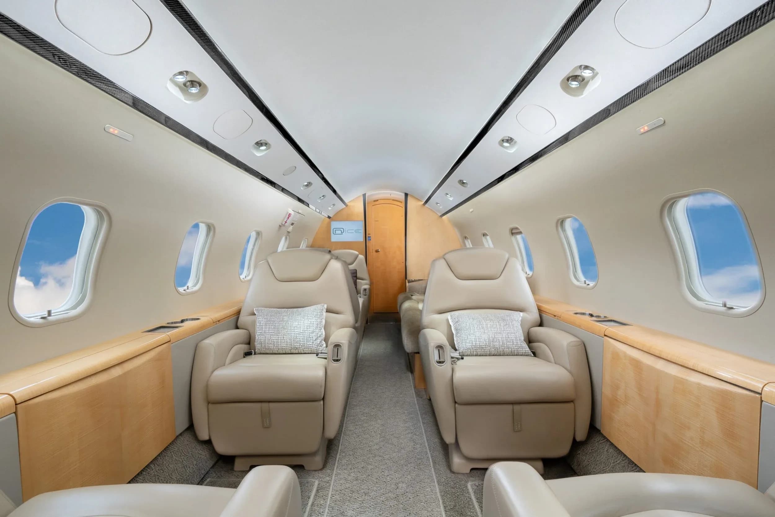 The interior of a private jet cabin with light beige leather seats and light wood paneling, looking towards the rear, with pillows on the seats.