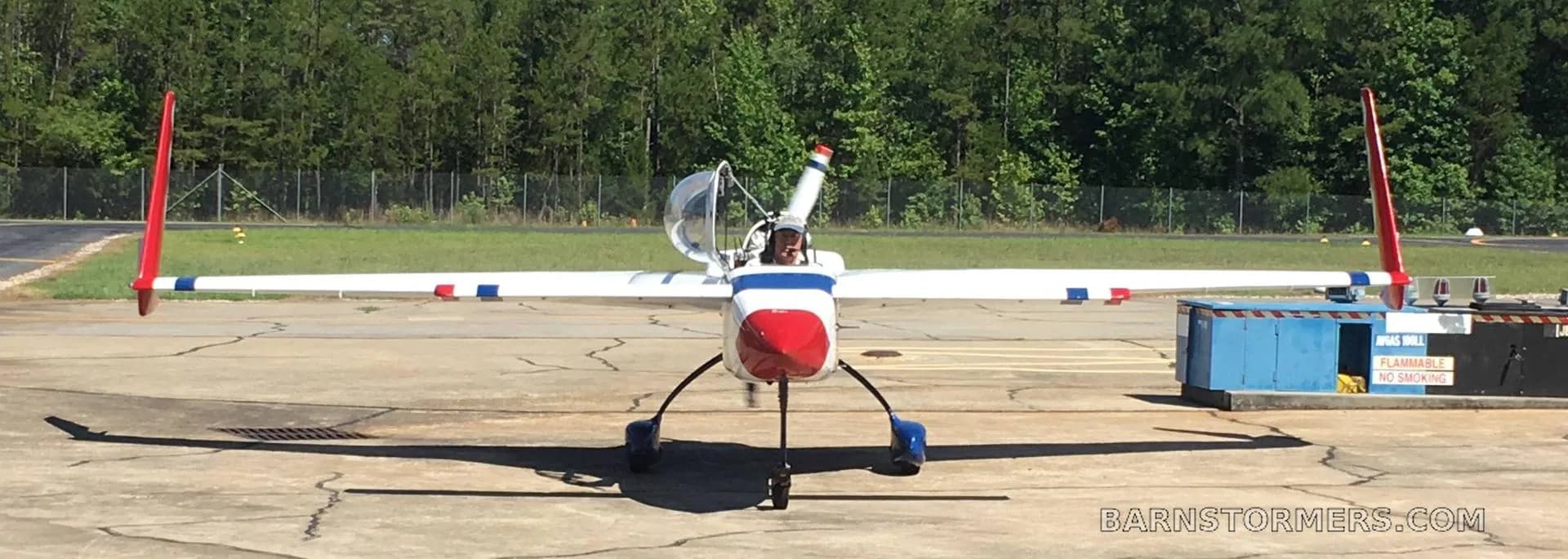 A white, red, and blue aircraft with twin tails and a pusher propeller, parked on an asphalt ramp with trees in the background.