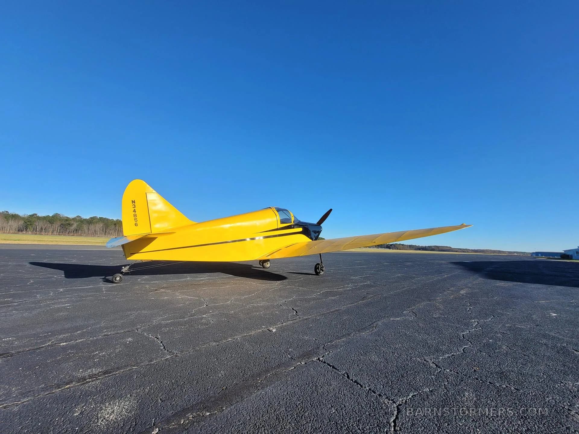 A yellow single-engine aircraft with black accents, viewed from the left side, on an asphalt runway with trees in the background.