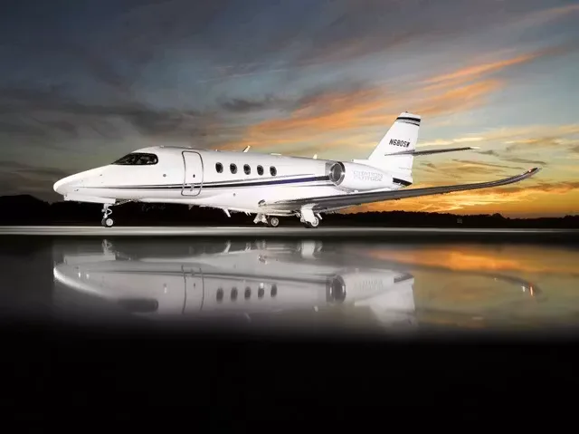 A white business jet with blue and grey stripes parked on a wet tarmac at sunset, reflecting in the water, viewed from the side.