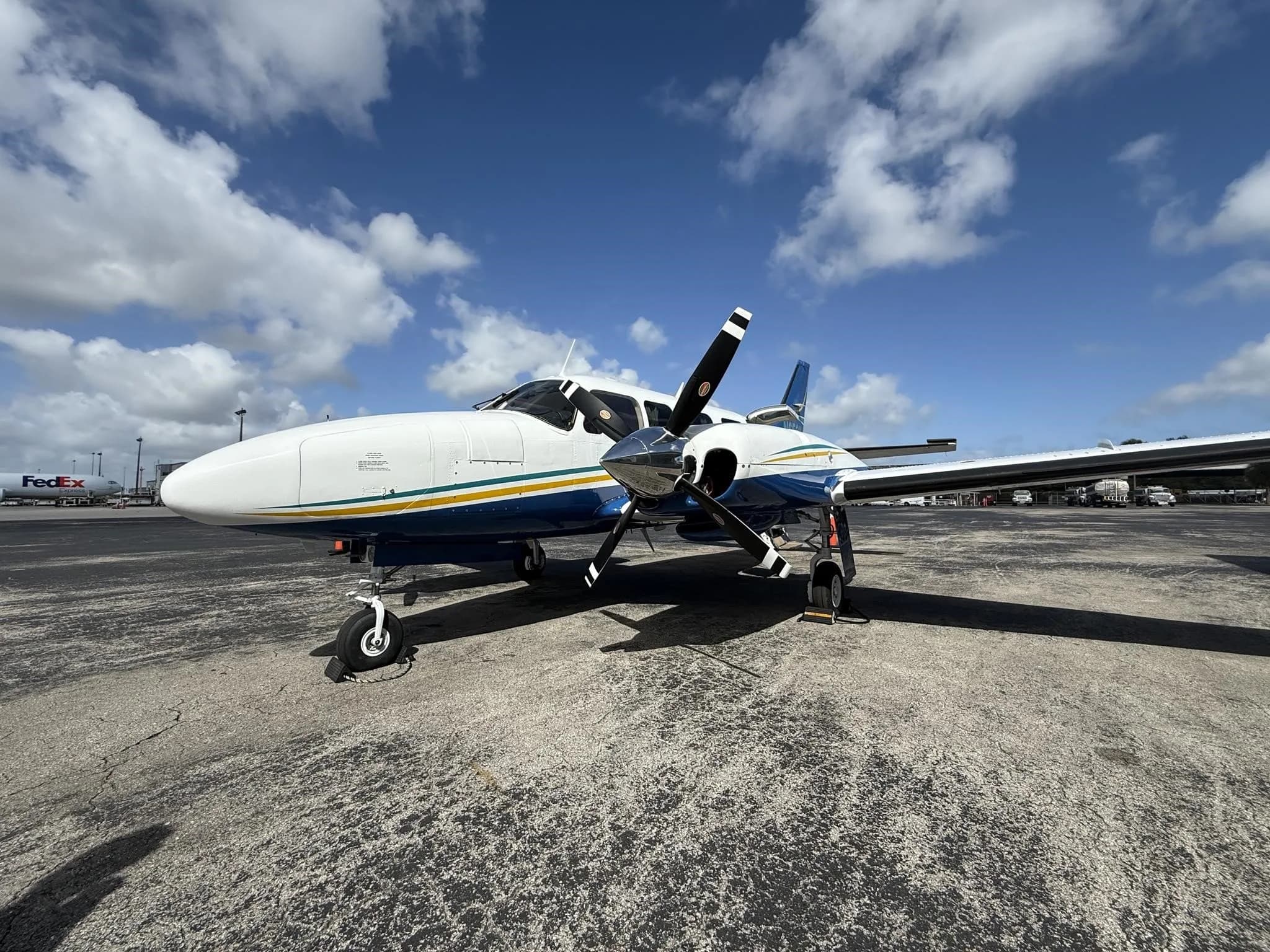 A white and blue twin-engine propeller aircraft with yellow and green stripes on the fuselage, parked on an airport tarmac under a partly cloudy sky.