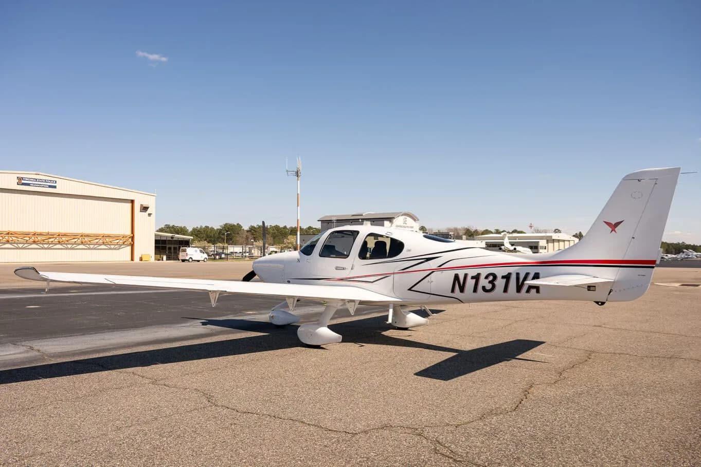 A white Cirrus SR22 aircraft with red and black accent stripes, tail number N131VA, parked on an airfield with a hangar in the background.