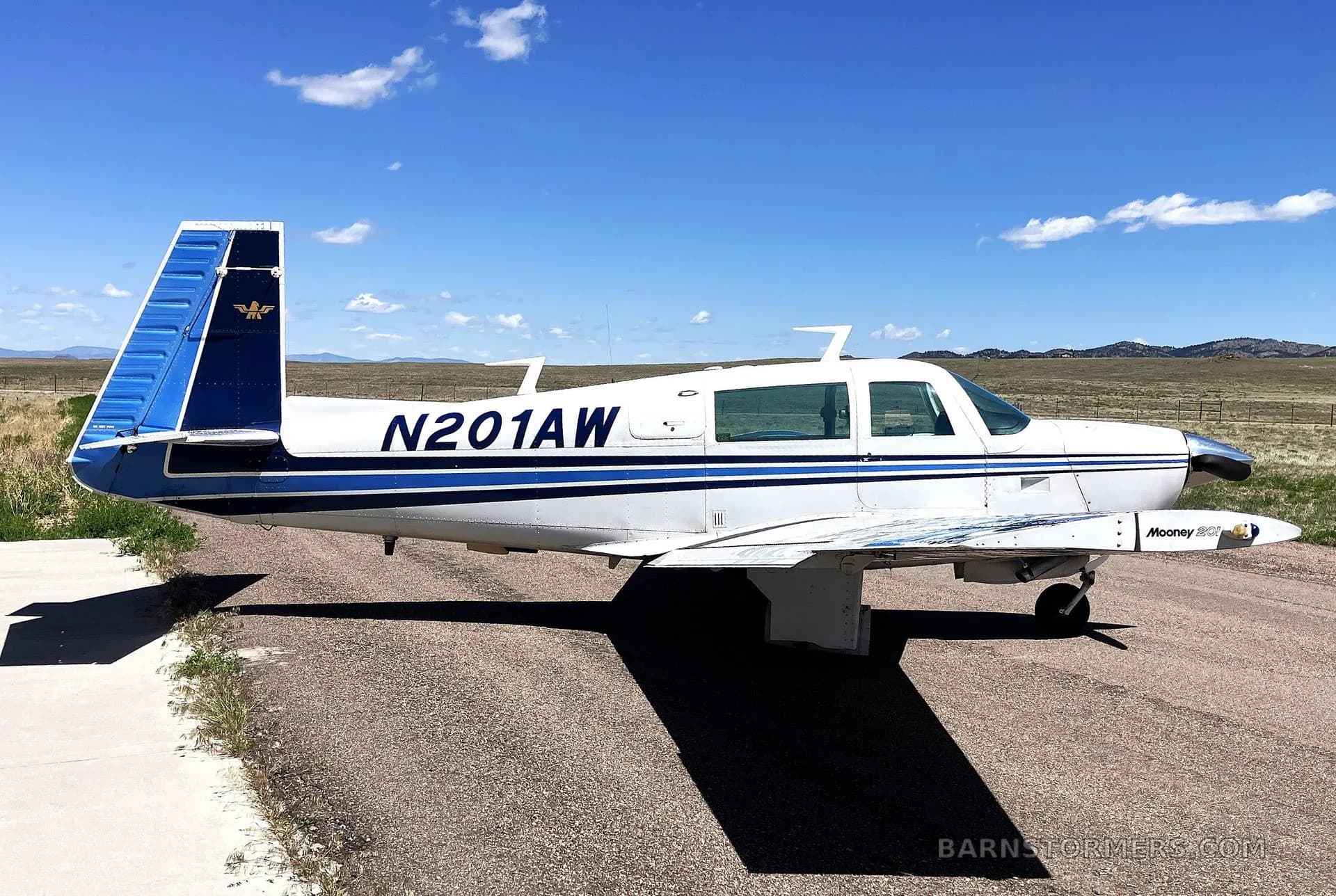 A white Mooney M20J aircraft with blue stripes and tail, registration N201AW, parked on an asphalt runway under a clear blue sky.