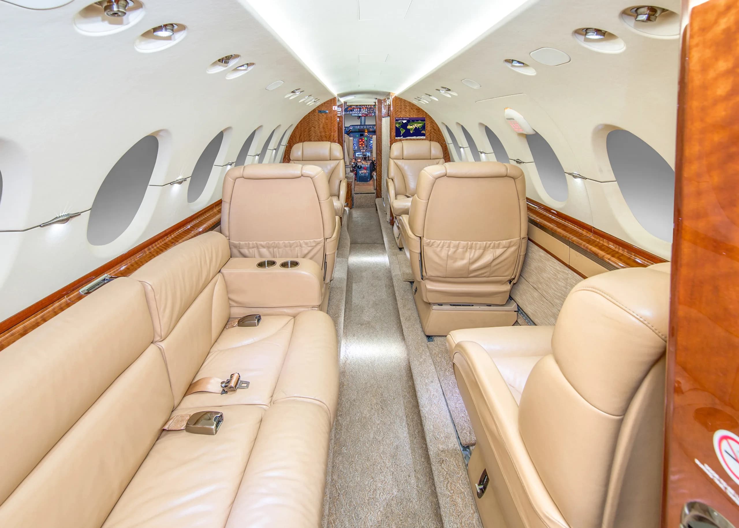 The spacious interior of a private jet cabin featuring a long tan leather sofa, individual seats, and wooden accents.