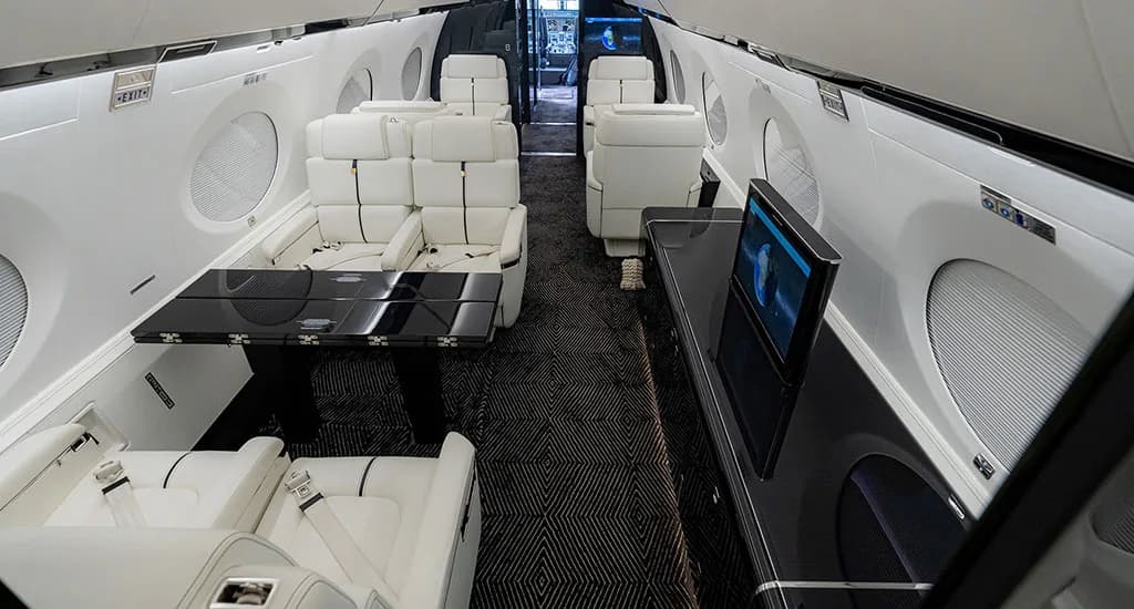 A long view of the business jet's elegant cabin interior with white seating, a black table, and a TV screen.