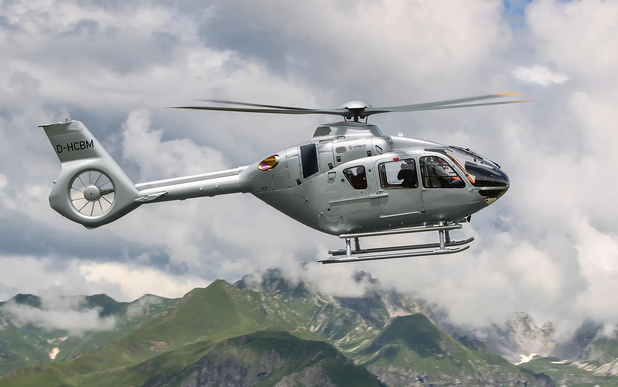 A silver Airbus H135 helicopter (D-HCBM) flying against a cloudy, mountainous backdrop.