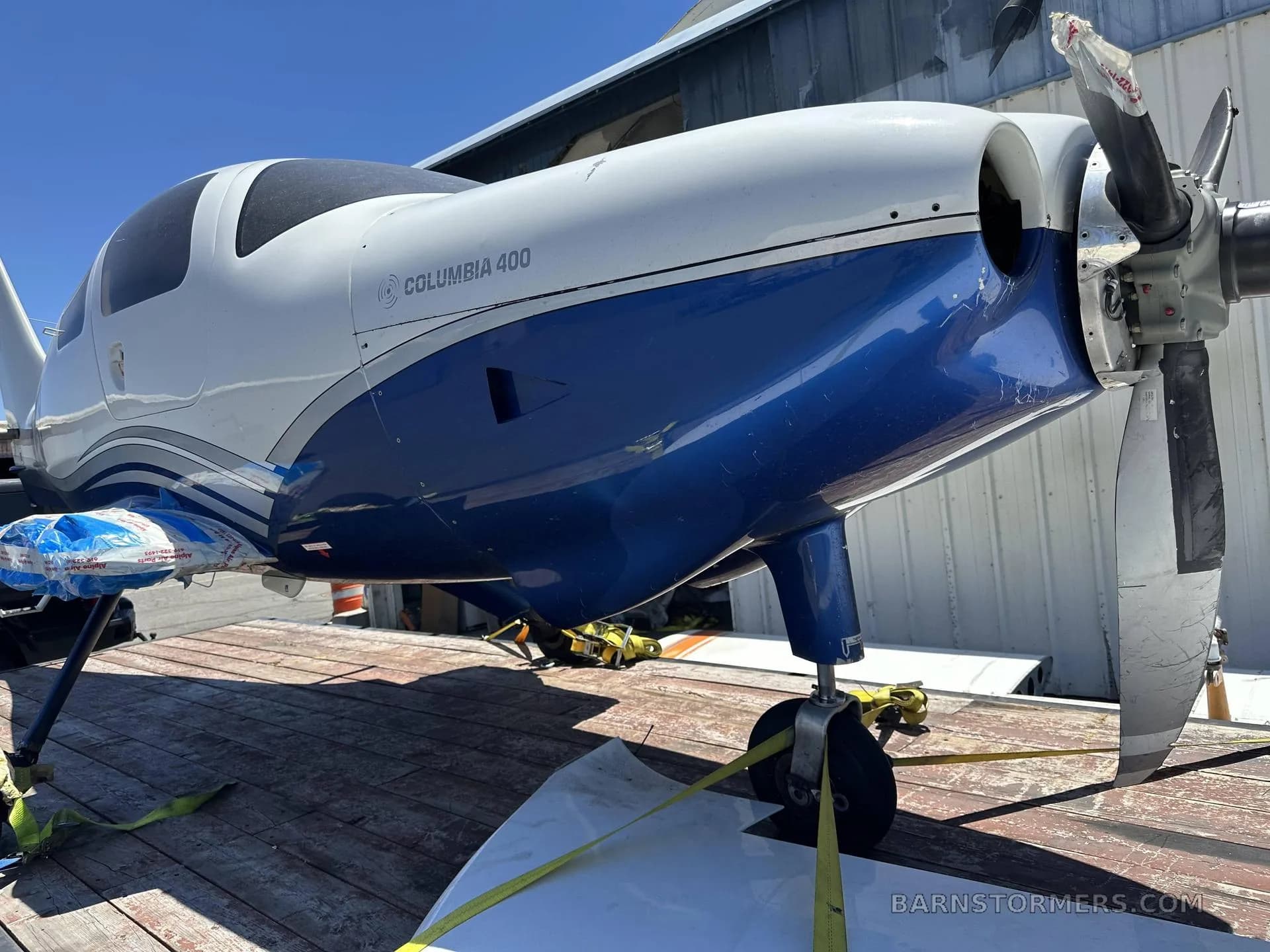 A front-side view of a white and blue Columbia 400 aircraft with a damaged propeller, secured on a wooden flatbed trailer.