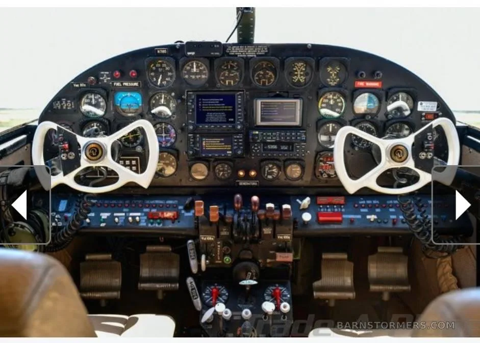 The cockpit of an aircraft featuring a complex instrument panel with gauges, screens, and two white control yokes.