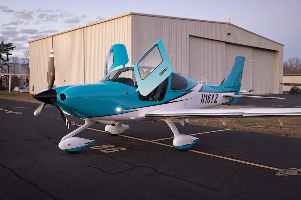 A turquoise and white Cirrus SR22 aircraft with its doors open, parked on an airfield in front of a hangar.
