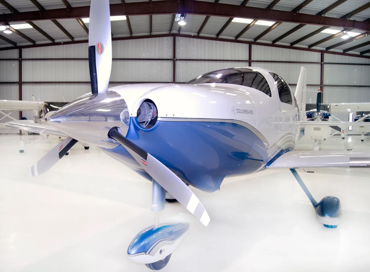 A close-up front view of a white and blue Columbia 400 aircraft with its propeller blades positioned vertically, inside a hangar.