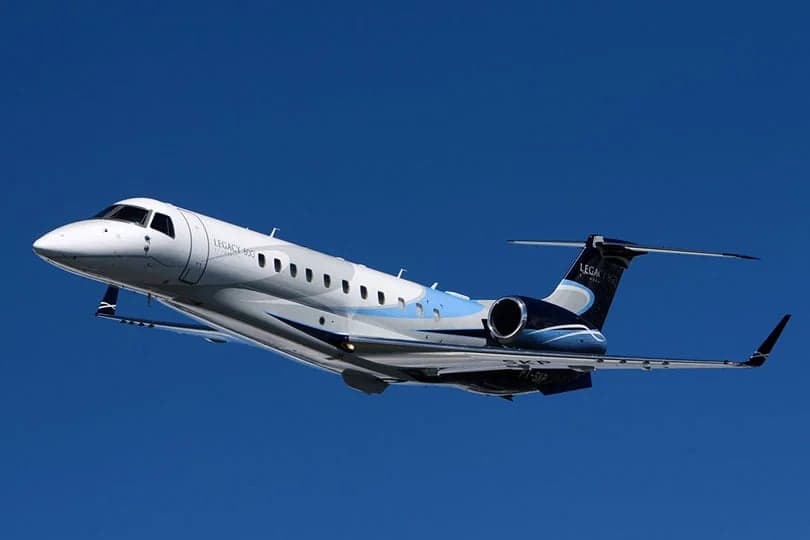 A white and blue Embraer Legacy 450 business jet with registration PR-SXP flies against a clear blue sky.