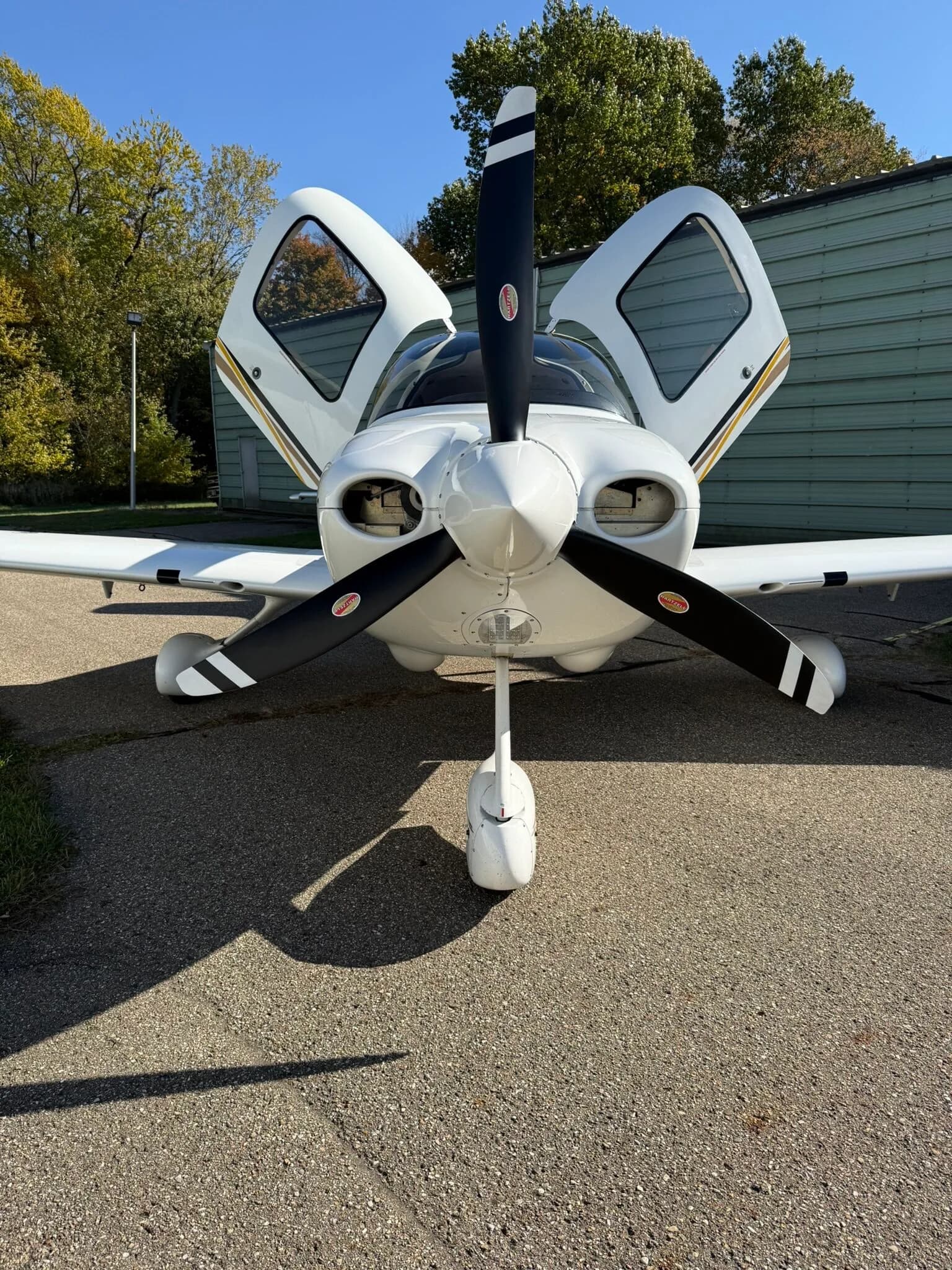 A head-on view of a white Cirrus SR20 aircraft with its propeller and open gull-wing doors visible, parked on an asphalt ramp.