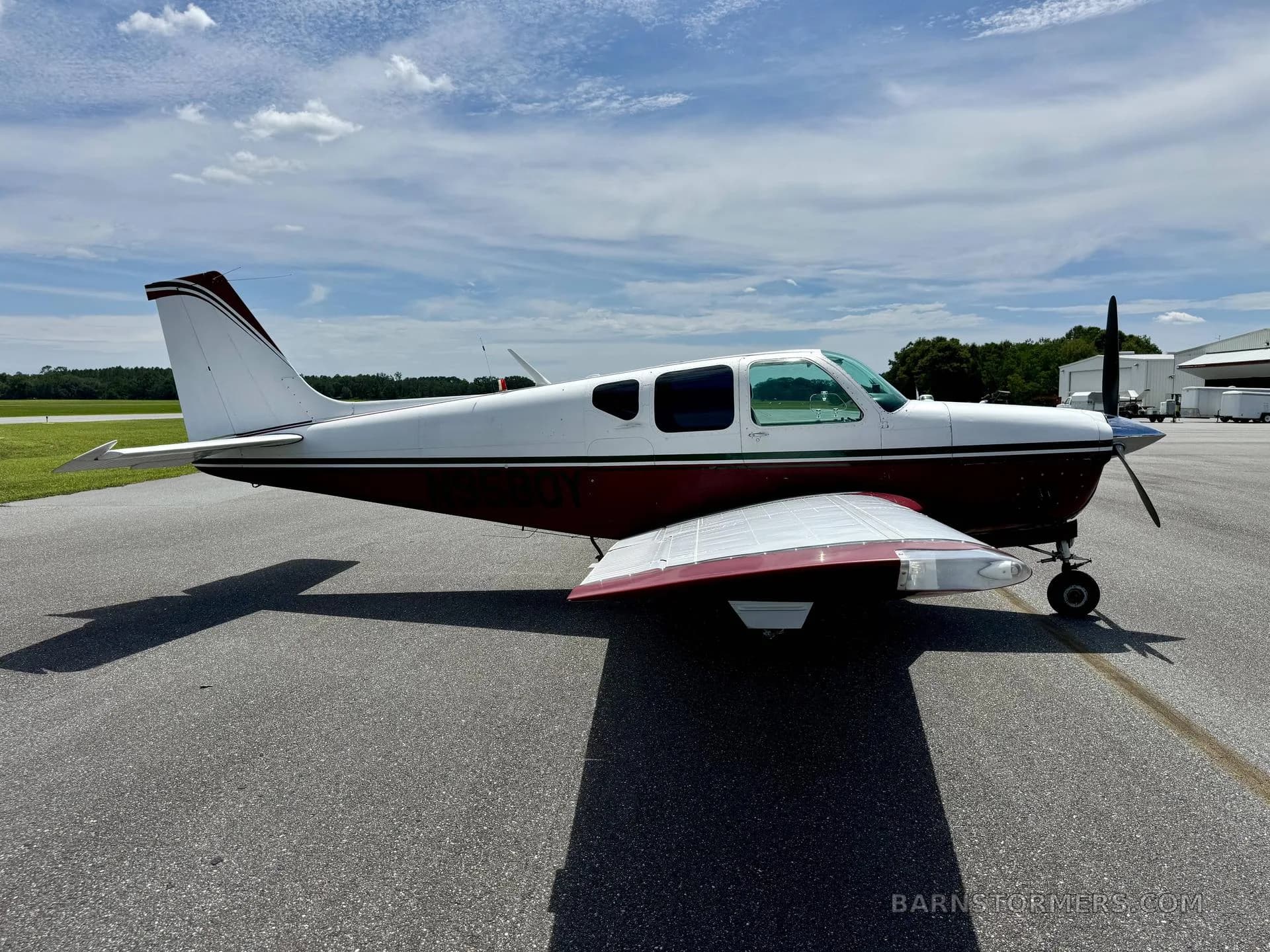 A white and maroon single-engine propeller aircraft is viewed from the side on an airfield under a sunny sky.