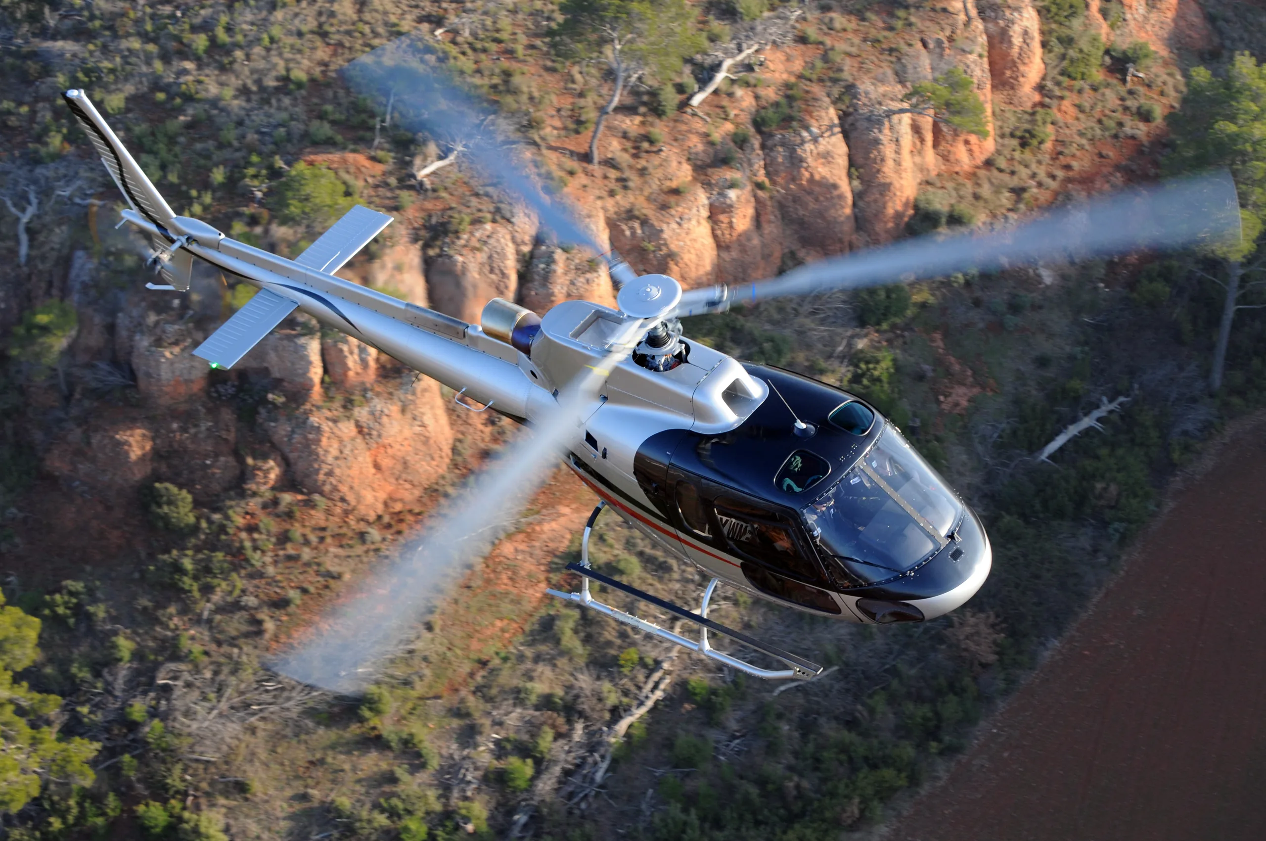A silver and black helicopter flies over a forest and canyon landscape.