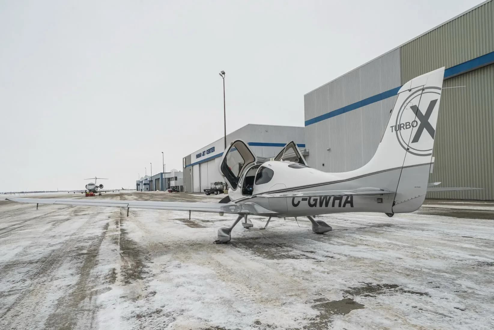 A white and grey Cirrus SR22T aircraft (C-GWHA) with its doors open, parked on a snowy tarmac with hangars and other aircraft in the background.