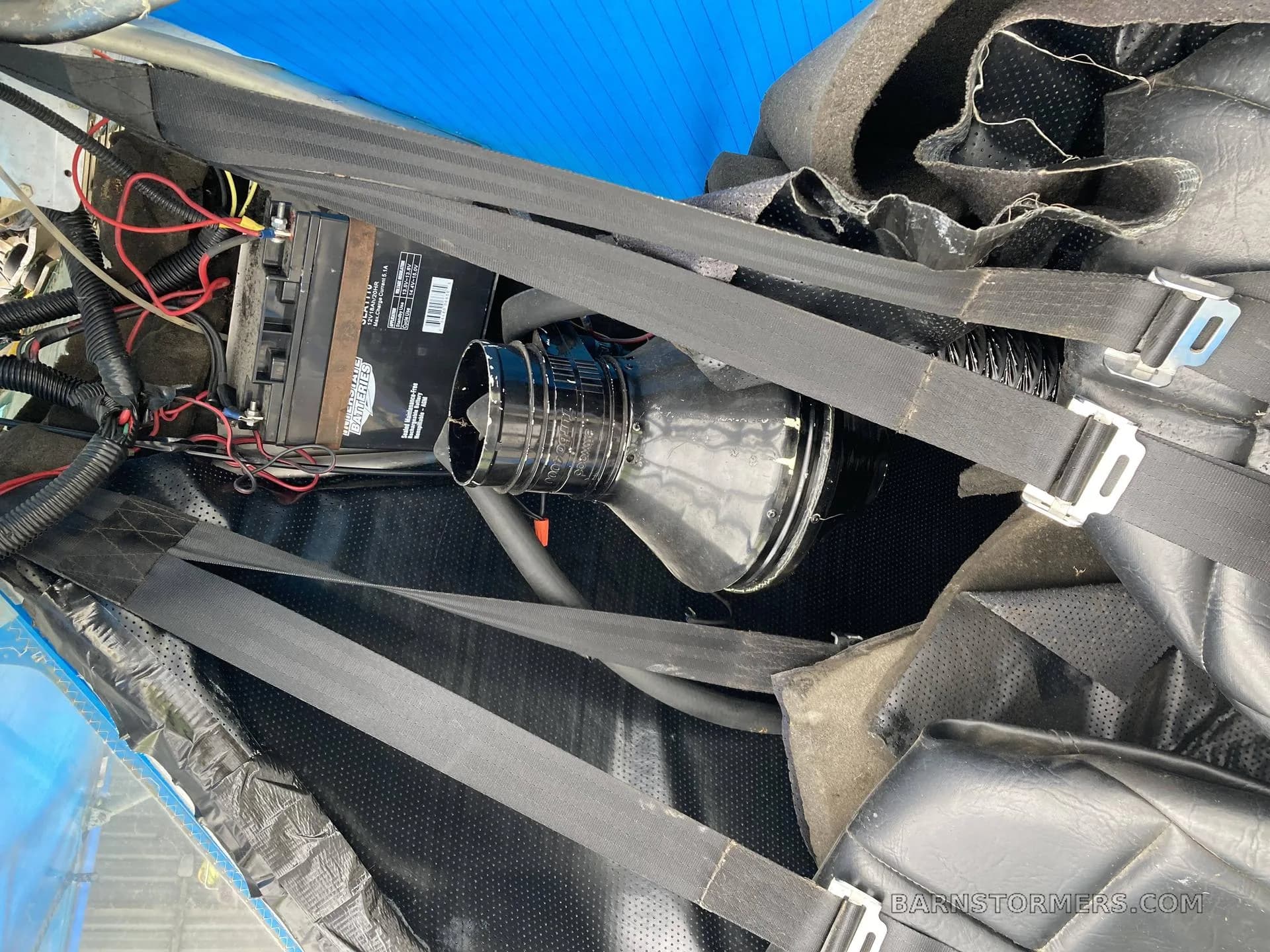 An overhead view of the aircraft's interior, showing a battery, wiring, and seatbelts in a black compartment.