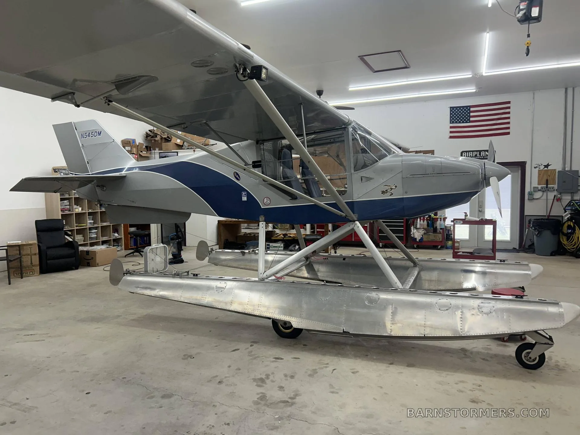 A silver and blue floatplane with registration N545DM is parked in a hangar, viewed from the rear-left.