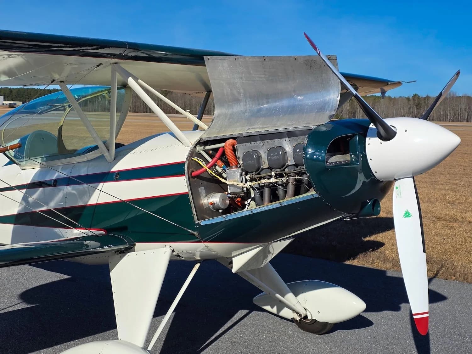 The engine cowling of the green and white biplane is open, revealing the engine components.