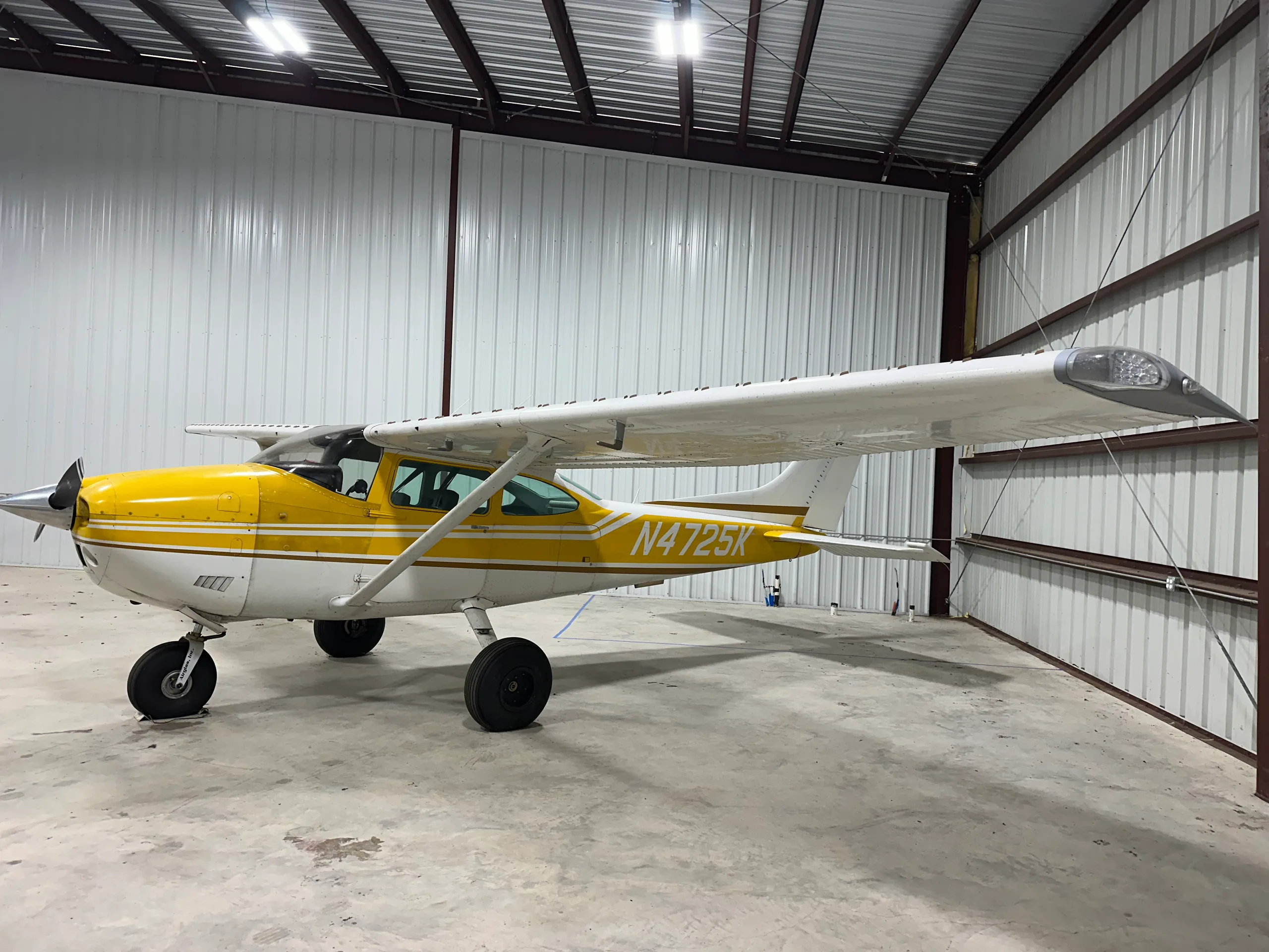 A yellow and white Cessna 172, N4725K, parked in a hangar, viewed from the front-left side.