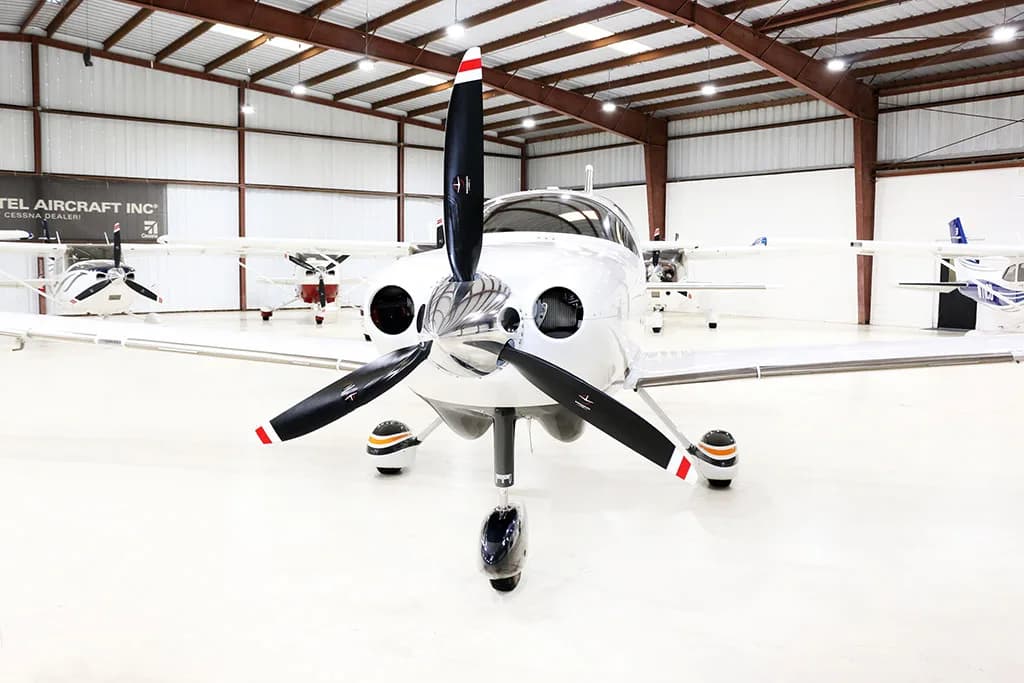 Front view of a white, black, and orange Cessna TTX aircraft with a three-bladed propeller, inside a hangar.