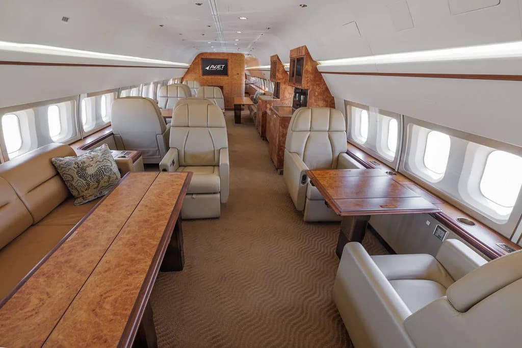 A wide view of the aircraft's luxurious interior cabin, featuring a long sofa, individual beige seats, and wooden tables.