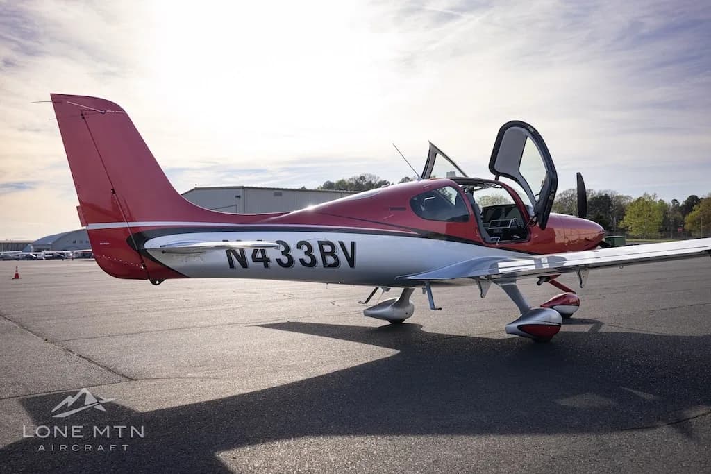 A side view of the red and silver Cirrus SR22 aircraft N433BV with its doors open, parked on an airfield.
