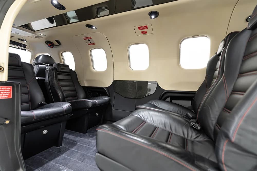 A side view of a small aircraft cabin showing black leather seats with red stitching and windows.
