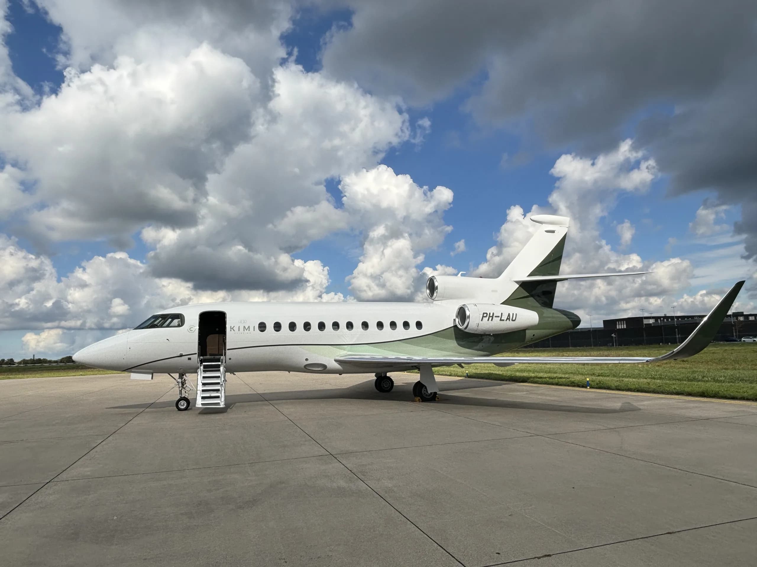 A white business jet with green accents on the tail and engines, registration PH-LAU, parked on an airfield under a cloudy sky.