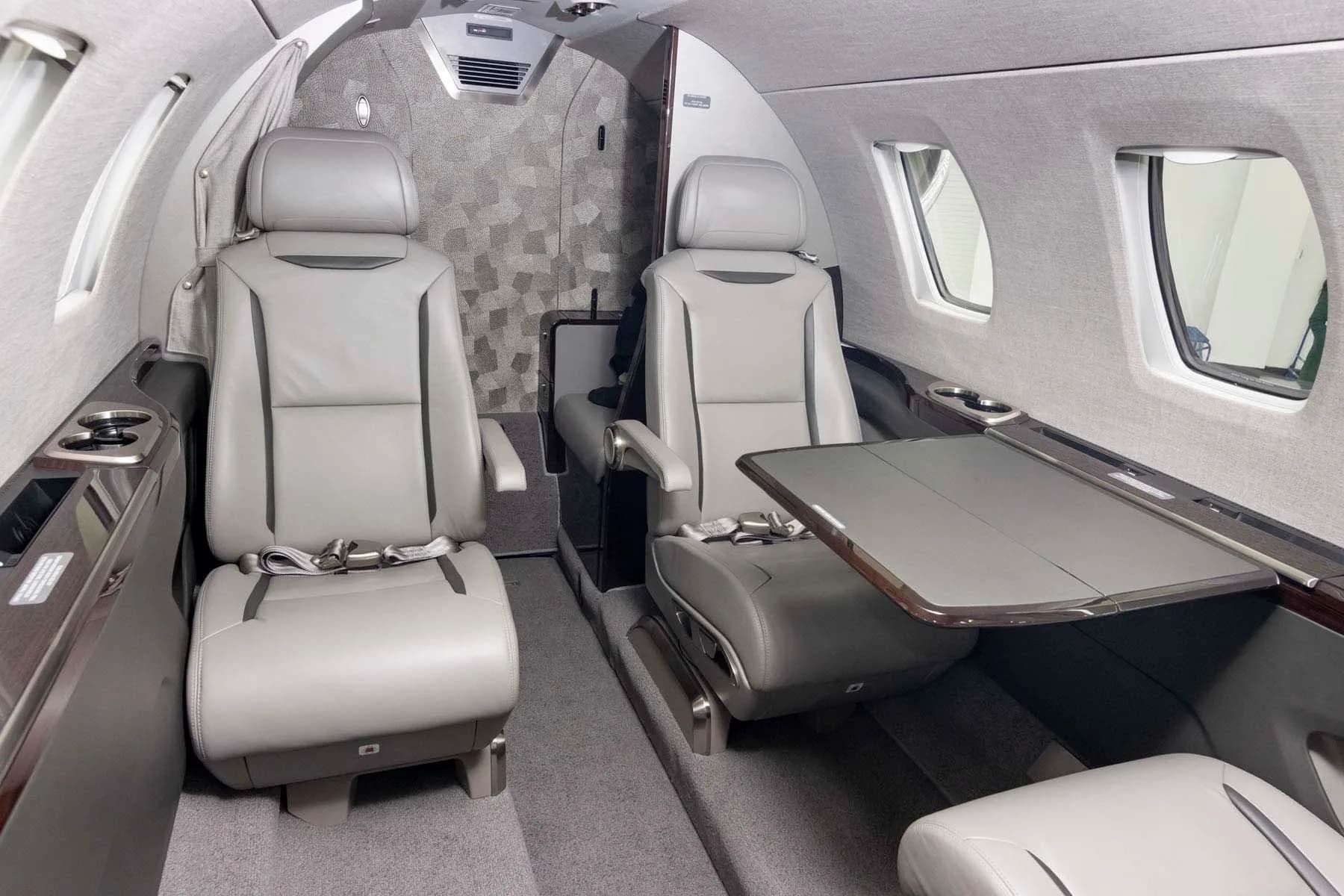 The interior cabin of a private jet, showing two grey leather passenger seats and a deployed table.