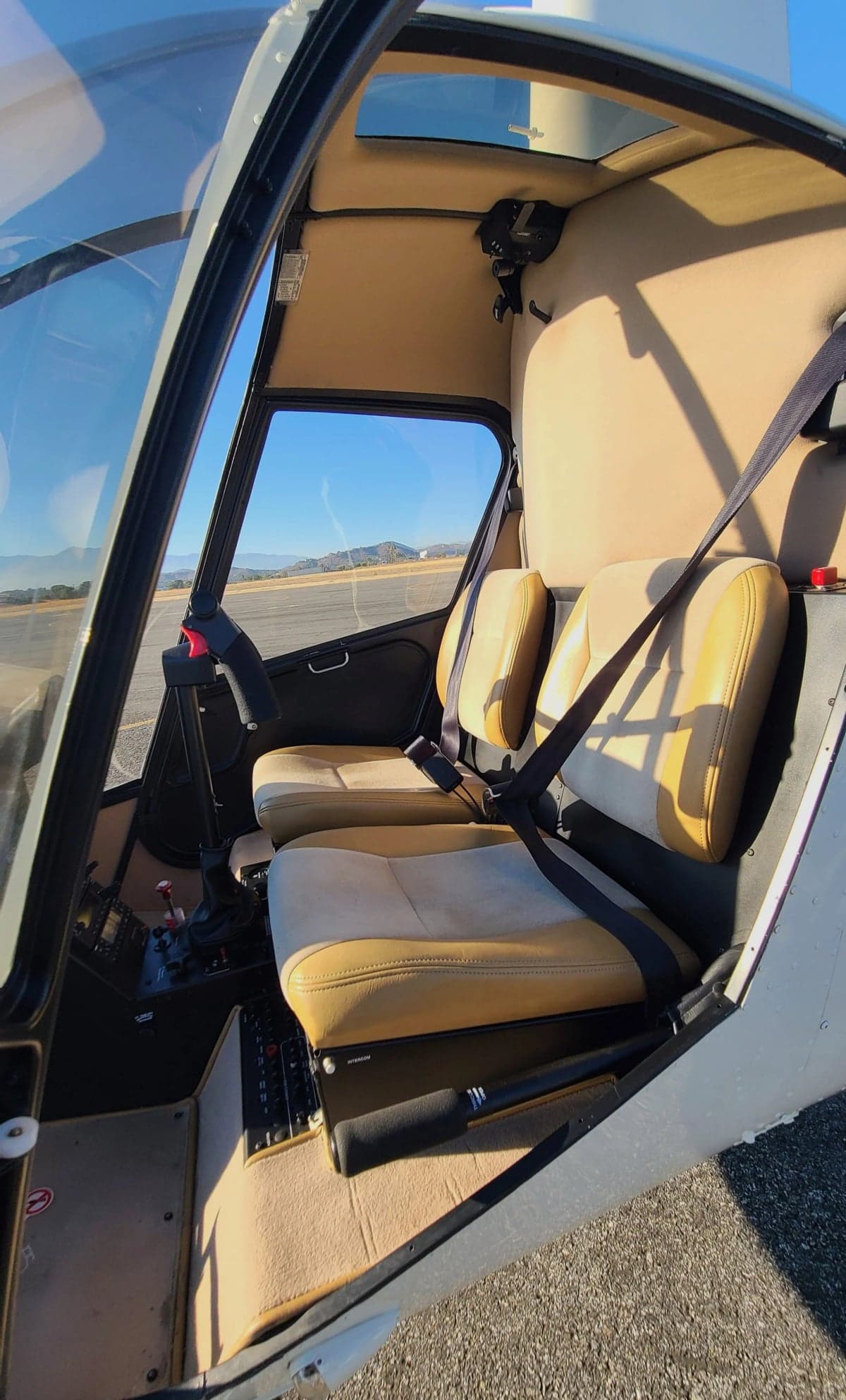 The tan interior seats and some controls of a Robinson R22 helicopter, viewed through the open door.