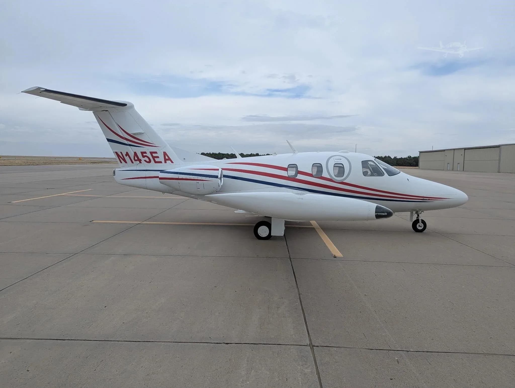 A white Eclipse jet with red and blue stripes, registration N145EA, parked on an airfield with a hangar in the background.