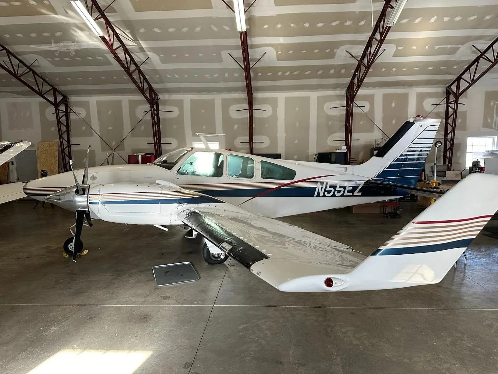 A white and blue twin-engine aircraft, N55EZ, is parked inside a hangar, viewed from the rear-left.