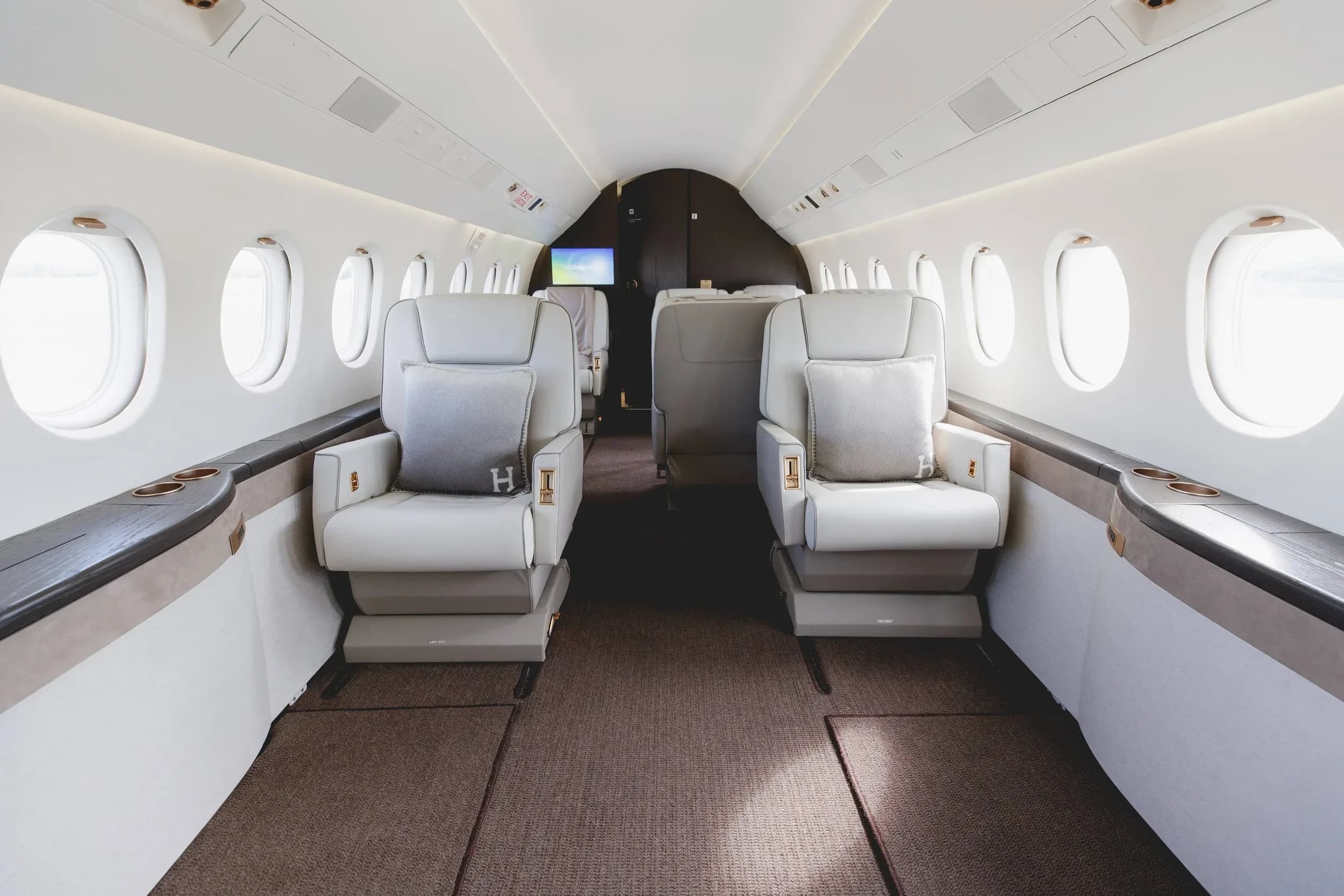 The luxurious interior cabin of a private jet, featuring two rows of light-colored leather seats with "H" monogrammed pillows.
