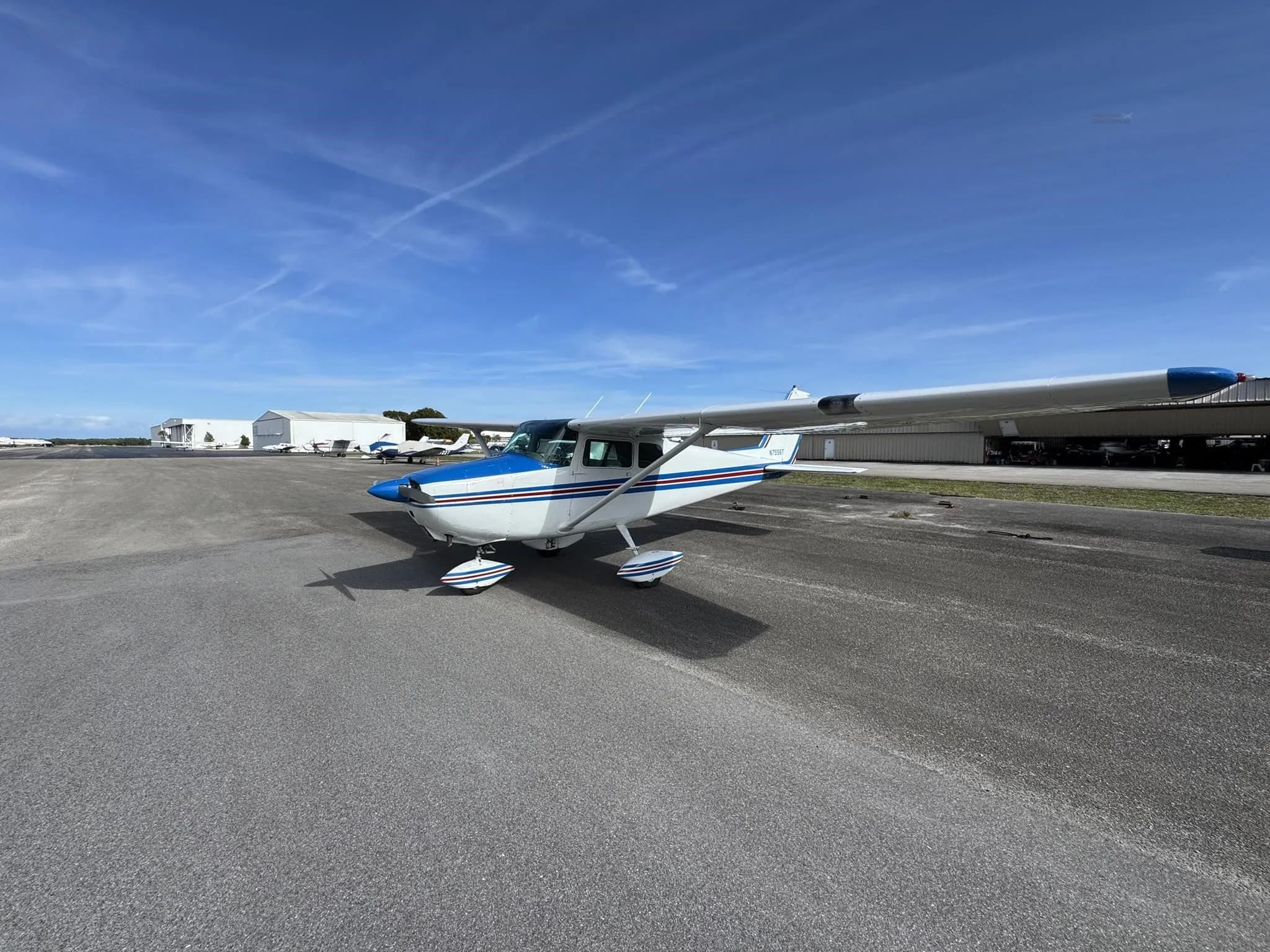 A white and blue Cessna 172 aircraft parked on a wide asphalt apron under a clear blue sky.