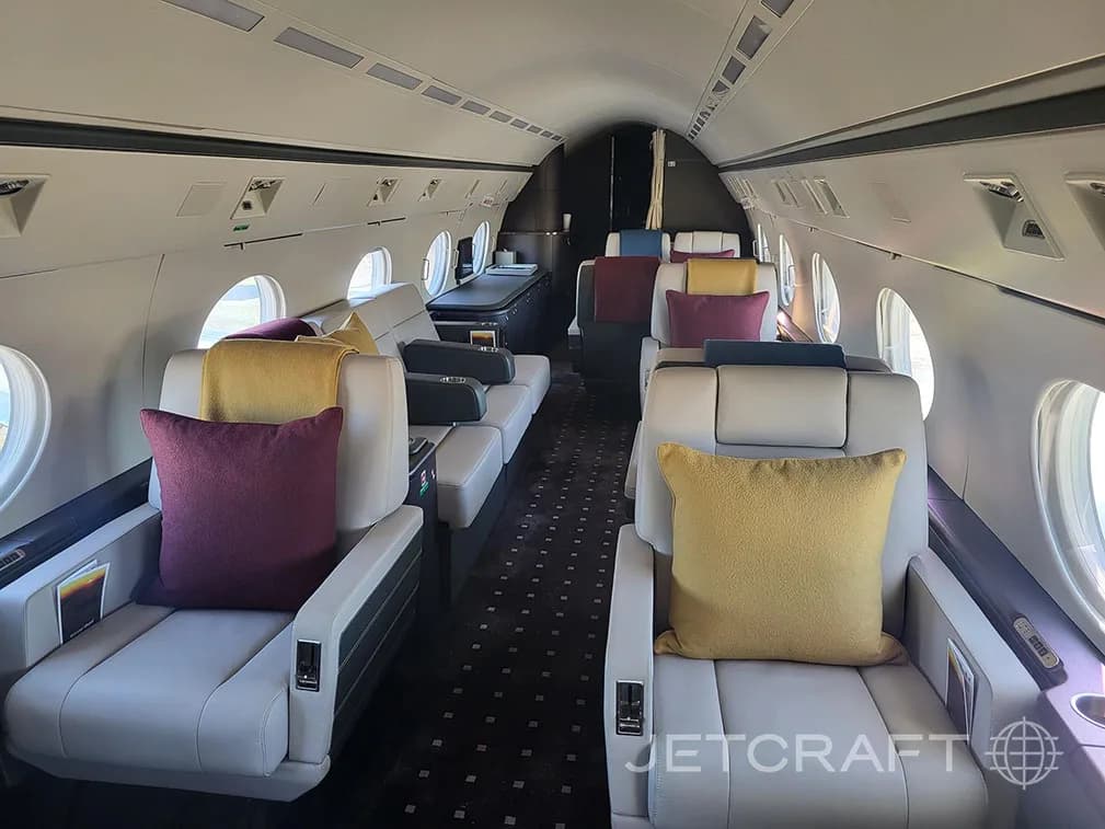 A wide view of the business jet cabin with light grey seats, colorful pillows, and large windows.