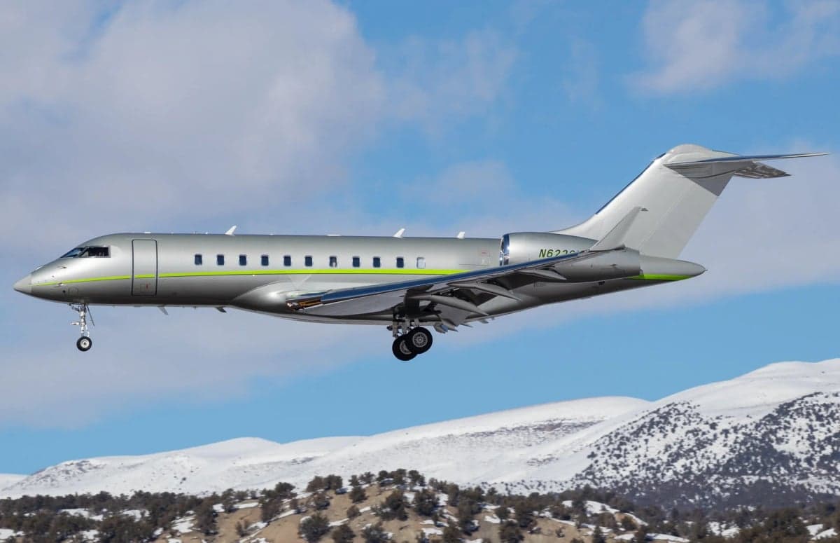 A Bombardier Global Express jet in flight over a snowy mountain landscape.