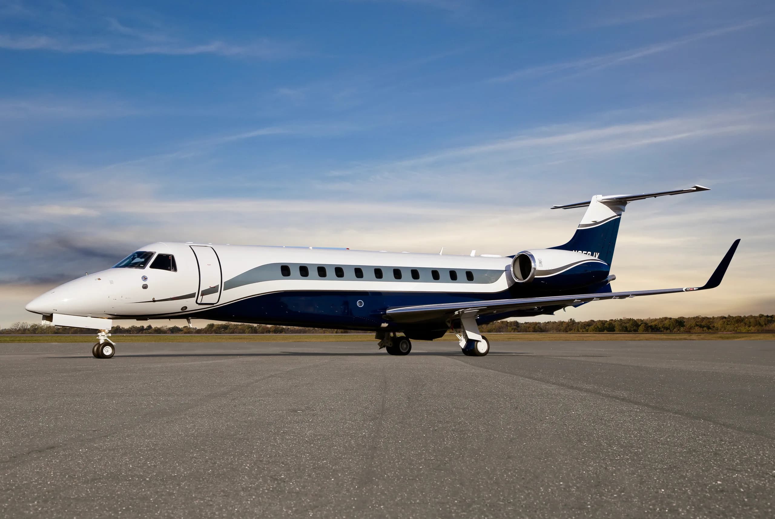 A dark blue, white, and grey Embraer Legacy business jet parked on an airfield under a partly cloudy sky.