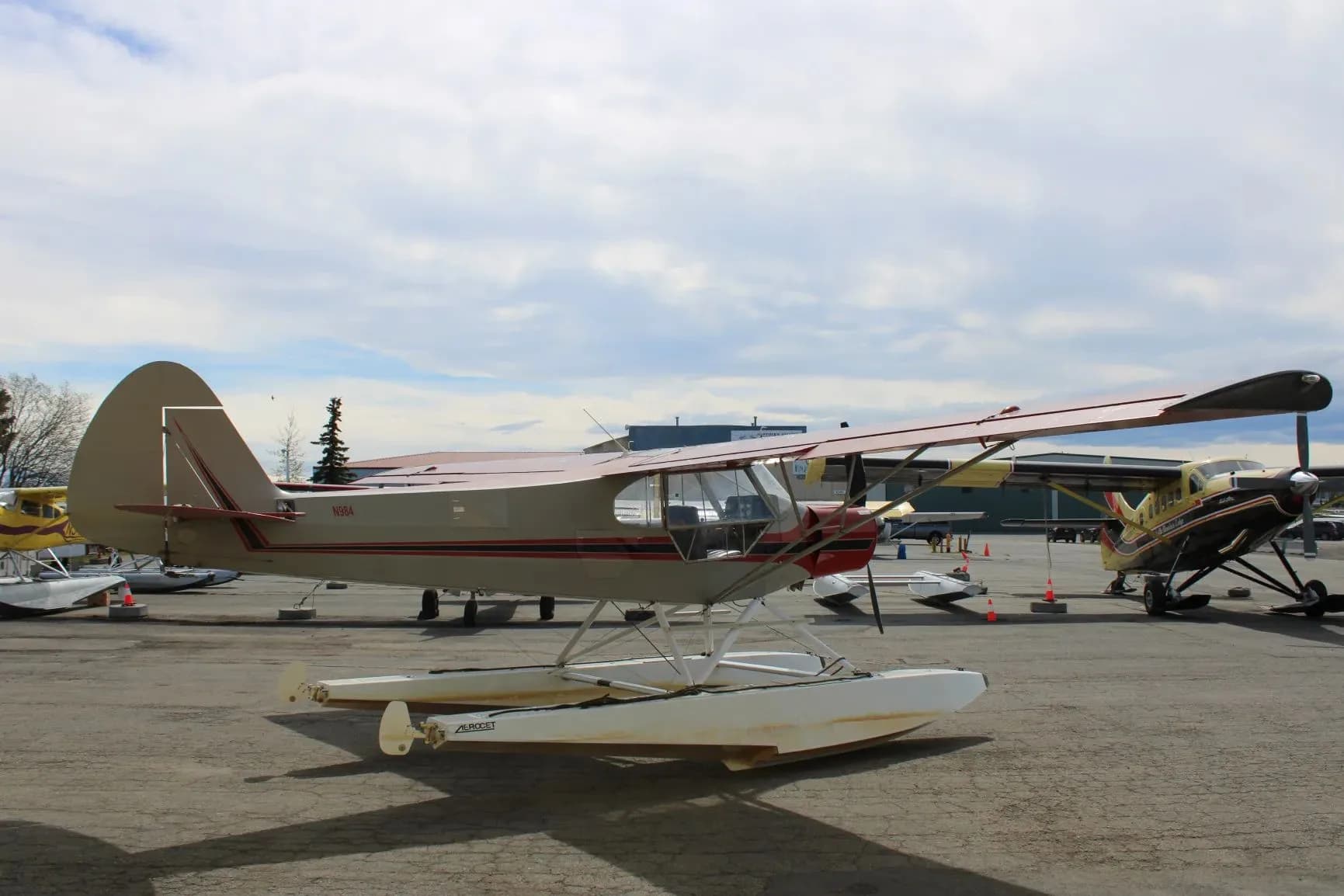 A side view of a red and grey floatplane with N984 on the tail, parked on an apron under a cloudy sky.