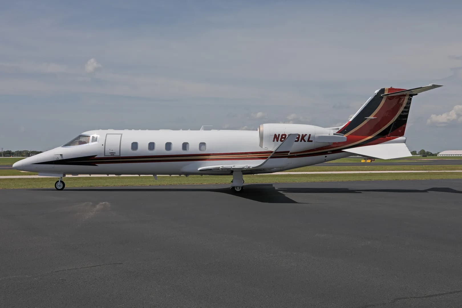 A white business jet with red and black tail accents, registration N873KL, parked on an asphalt runway under a partly cloudy sky.