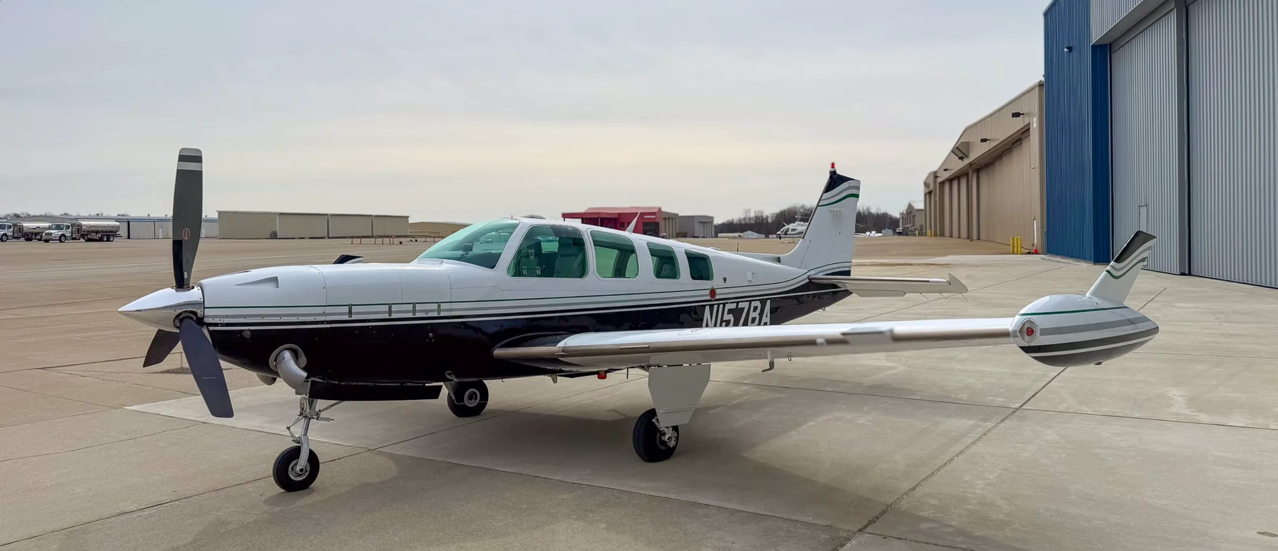 A three-quarter front view of the white and black Beechcraft Baron N157BA on an airfield, with other buildings in the distance.