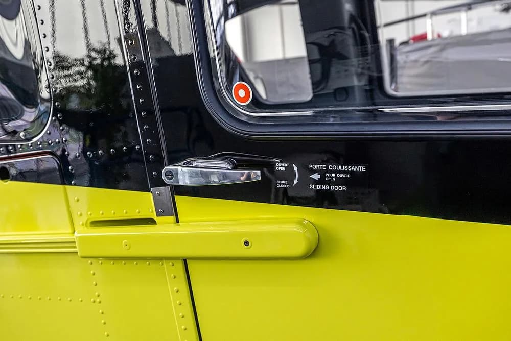 A close-up of the bright yellow and black side of an Airbus H135 helicopter, showing a sliding door handle and instructions.